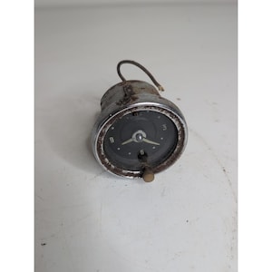 May include: A classic car clock with a dark face, displaying white numerals at the 3, 6, and 9 o'clock positions. It has a chrome bezel and a brass winding knob. A wire protrudes from the top of the clock.