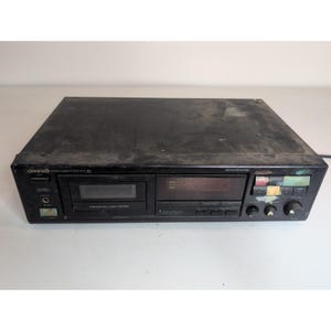 May include: A black Onkyo stereo cassette tape deck featuring a central cassette compartment, a digital display, multiple control buttons, and a headphone jack. The unit has a rectangular chassis with visible dust on the top surface and front panel.