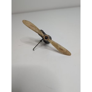 Puede incluir: Una hélice de avión modelo vintage con dos palas. Las palas son de color beige descolorido, y el cubo central es negro con intrincados detalles mecánicos. Un pequeño soporte metálico sostiene la hélice.