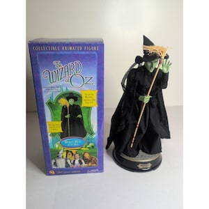 Può includere: Una statuetta animata da collezione della Strega dell'Ovest dal Mago di Oz, con pelle verde, abito nero e cappello a punta. La figura tiene in mano una scopa e si trova su una base circolare accanto alla sua scatola blu originale.
