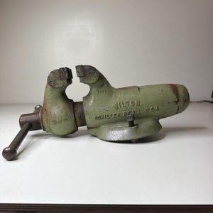 May include: A vintage light green metal bench vise with a dark brown handle. The body features the embossed text WILTON and SCHILLER PK ILL. U.S.A. The tool has a heavy-duty construction with a wide base and a screw mechanism for clamping objects.