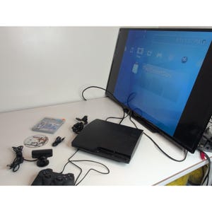 May include: A black PlayStation 3 game console connected to a flat-screen television displaying the PlayStation Store interface. Included are a black game controller, a small black camera, a game disc, a game case, and various black power and data cables.