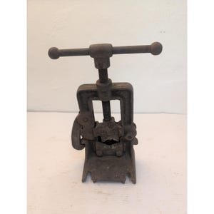 May include: A dark gray, weathered cast iron pipe cutter tool featuring a vertical threaded screw mechanism, a horizontal handle with rounded ends, and a central jaw assembly designed for gripping and cutting cylindrical metal pipes.