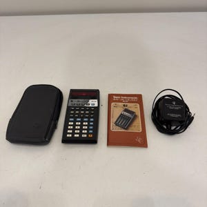 May include: A black Texas Instruments SR-50 slide rule calculator with a numeric keypad, a black carrying case, a brown instructional manual titled Texas Instruments slide rule calculator SR-50, and a black power adapter with a coiled cord.
