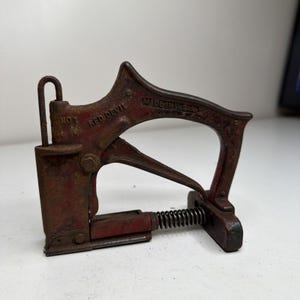 Puede incluir: Grapadora vintage Red Devil No. 1. La herramienta es de metal y tiene un acabado rojo con signos de desgaste. Las palabras "RED DEVIL" y "MADE IN USA" son visibles en la herramienta.