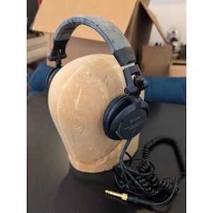 Puede incluir: Auriculares estéreo negros Sony MDR-V600 con una diadema gris ancha y desgastada, bisagras ajustables y un cable negro en espiral con un conector jack de 6,35 mm chapado en oro. Las copas circulares muestran la marca Sony y el modelo.