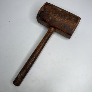 Könnte beinhalten: Ein hölzerner Vintage-Schlägel mit rechteckigem Kopf und zylindrischem Stiel. Das Holz weist eine dunkelbraune, verwitterte Oberfläche mit sichtbarer Maserung, oberflächlichen Kratzern und Gebrauchsspuren am gesamten Werkzeug auf.