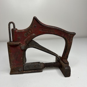 May include: A vintage red and black metal staple gun, featuring the text "RED DEVIL" and "NO. 1". The tool has a spring and a curved handle. This staple gun is likely used for construction or upholstery, and is a classic tool.