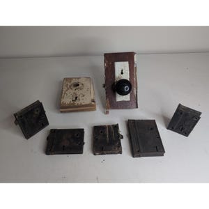 May include: A collection of six vintage door hardware components including rectangular metal lock plates and a single door knob assembly. The set features weathered white and brown finishes on some plates, while others are solid black metal.