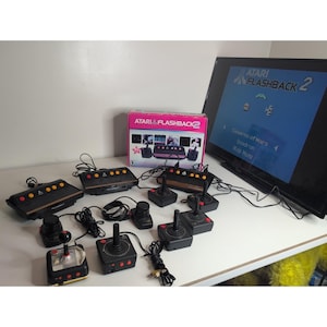 Puede incluir: Un conjunto de consolas de videojuegos Atari Flashback 2 que incluye tres consolas negras con detalles de madera, cinco joysticks negros con botones rojos y una caja rosa y blanca. Una pantalla de televisión muestra la interfaz del menú Atari.