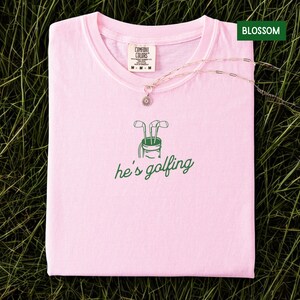 May include: A light pink t-shirt with the text "he's golfing" and a green graphic of golf clubs in a golf bag. A silver necklace with a pendant is draped across the shirt. The shirt has a tag that says "Comfort Colors".