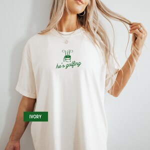 May include: Ivory-colored t-shirt with a green golf club graphic and the text "he's golfing". The shirt is made of a soft material. The word "IVORY" is displayed on a green rectangle.