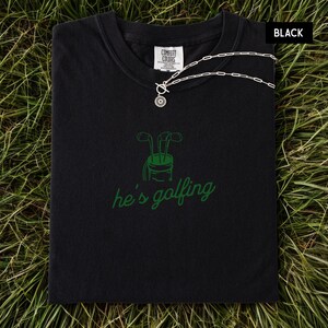 May include: Black t-shirt with a green golf club graphic and the phrase "he's golfing." A silver necklace with a circular pendant rests on the shirt. The shirt is folded on a bed of green grass.