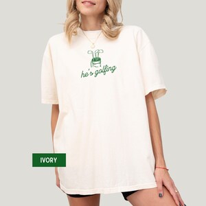 May include: Ivory-colored t-shirt with a green golf bag graphic and the text "he's golfing." The shirt is a relaxed fit. The word "IVORY" is in a green box at the bottom left.