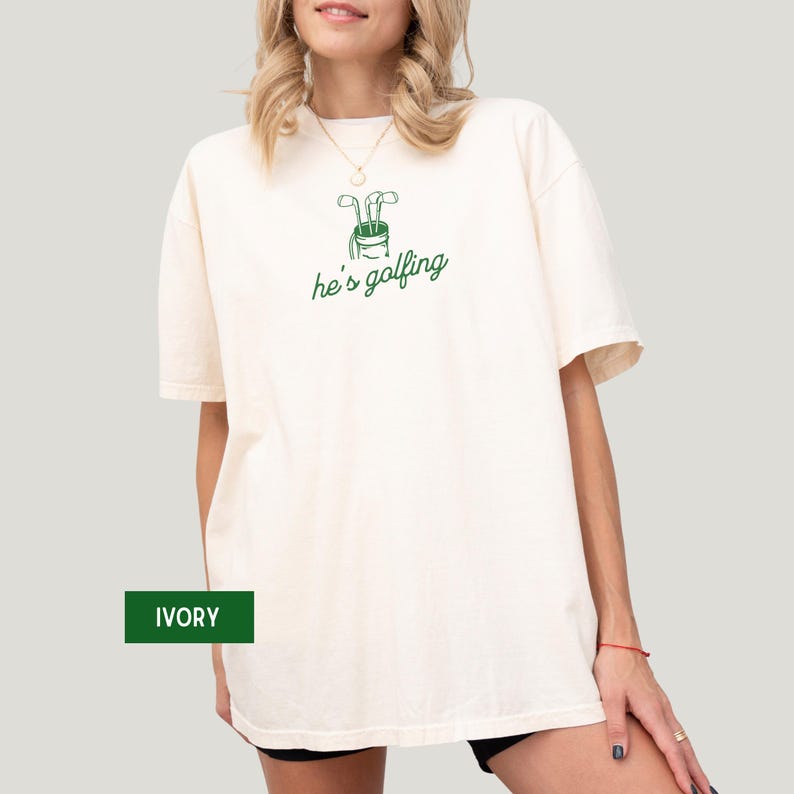 May include: Ivory-colored t-shirt with a green golf bag graphic and the text "he's golfing." The shirt is a relaxed fit. The word "IVORY" is in a green box at the bottom left.