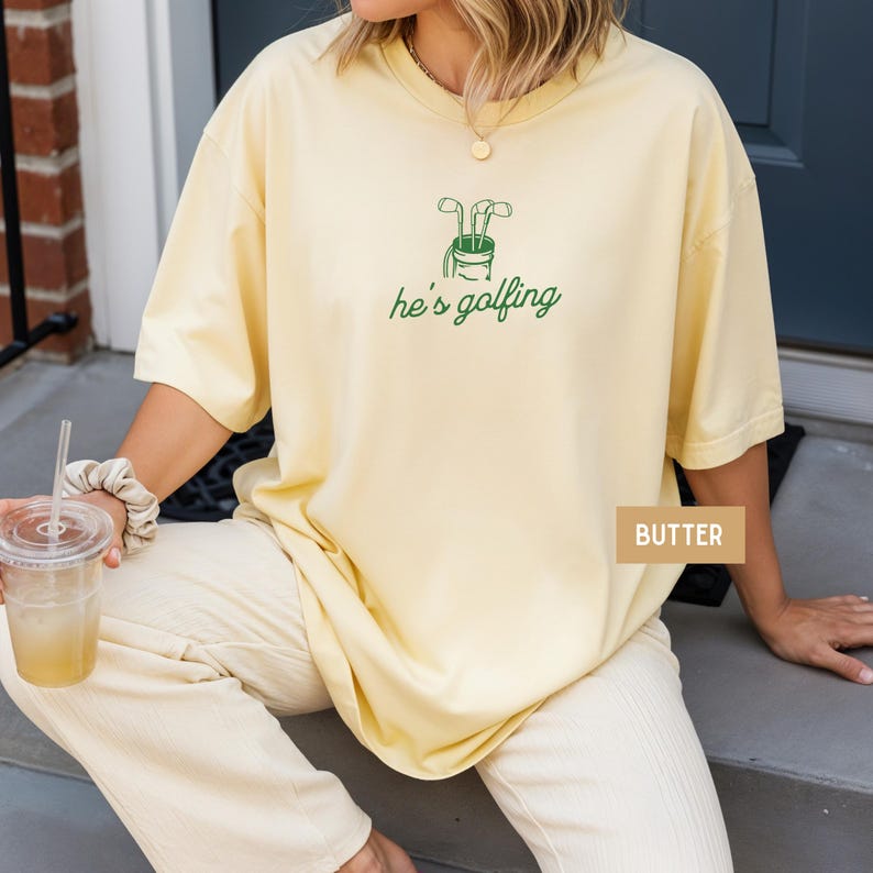 May include: Butter-colored t-shirt with a green golf club graphic and the text "he's golfing." The shirt has short sleeves and a relaxed fit. The word "BUTTER" is printed on a small rectangular label.