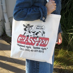 May include: A white canvas tote bag with a cartoon cow graphic and the words "GRASS-FED" in red. The bag also features the text "Raised right. Tastes right." and a phone number.