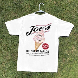 May include: White t-shirt with the text "Joe's" in a stylized font, above a cartoon ice cream cone with a smiling face. Below, the text reads "ICE CREAM PARLOR" and a tagline. The shirt is hanging on a white hanger.