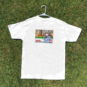 Peut inclure: T-shirt blanc avec un dessin animé. Le dessin représente un journaliste devant un bâtiment en feu, avec le texte "Fiery but Mostly Peaceful" en dessous. Le t-shirt est suspendu à un cintre blanc sur un fond d'herbe verte.