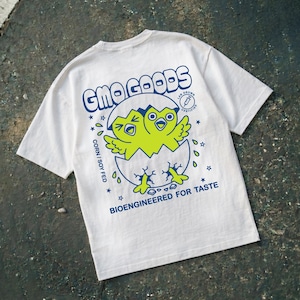 May include: A cream-colored short-sleeve t-shirt featuring a blue and lime green graphic on the back. The design shows a two-headed chick hatching from an egg with the text GMO GOODS, LAB GROWN CERTIFIED, CORN / SOY FED, and BIOENGINEERED FOR TASTE.
