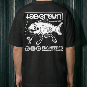 May include: A black short-sleeve t-shirt featuring a white graphic on the back. The design shows a fish with legs, a stick figure fisherman, and the text "LAB GROWN MODIFIED SALMON" and "BIO ENGINEERED MUTATED TASTE!" in a distressed, bold font.