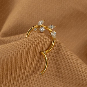 Puede incluir: Un pendiente de oreja dorado con un diseño de rama, adornado con piedras preciosas transparentes y brillantes. La pieza se curva alrededor de la oreja, sobre un fondo de tela marrón, creando una estética delicada y elegante.