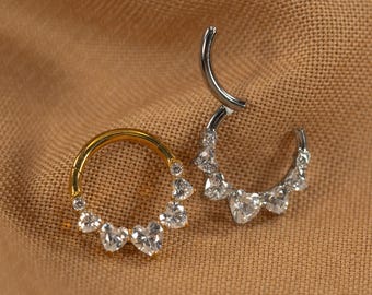 Pode incluir: Dois anéis de septo articulados com pedras preciosas transparentes em forma de coração. Um anel é dourado, o outro é prateado. As joias são exibidas em um fundo de tecido castanho.