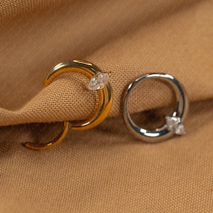 May include: Two rings, one gold and one silver, rest on a tan fabric. The gold ring has a crescent shape with a marquise-cut diamond. The silver ring is circular with three small diamonds.