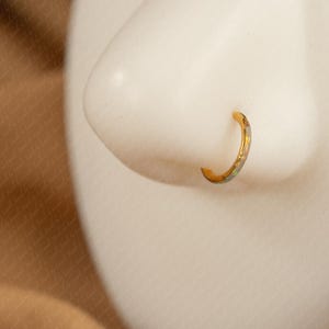 May include: A gold-colored nose ring with a subtle, patterned design. The hoop-style jewelry is shown on a white mannequin nose, set against a neutral background. The ring's design includes hints of silver and blue.