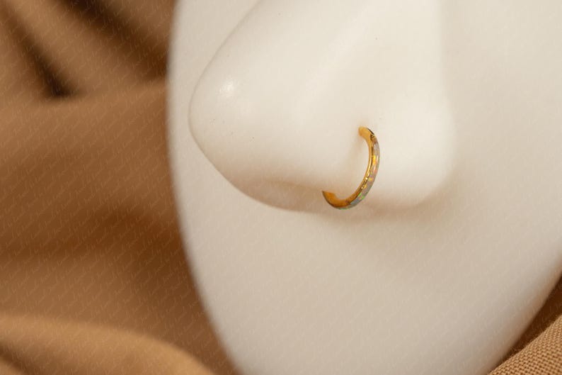 May include: A gold-colored nose ring with a subtle, patterned design. The hoop-style jewelry is shown on a white mannequin nose, set against a neutral background. The ring's design includes hints of silver and blue.