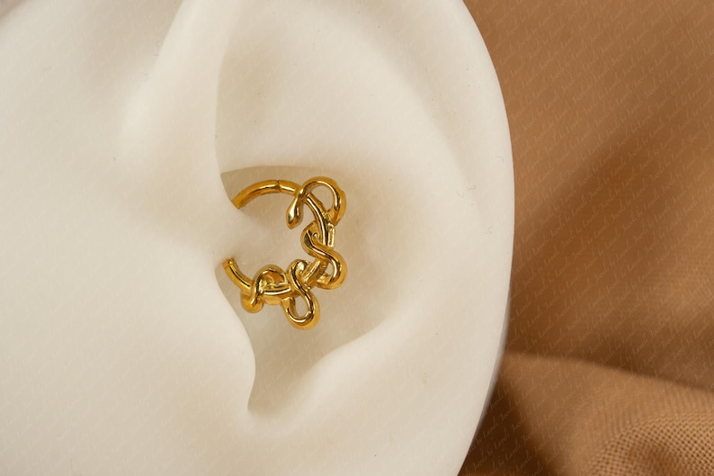 May include: A gold-colored, decorative ear piercing hoop. The hoop features a looped design and is set against a white background, with a tan background in the corner.