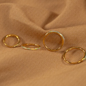 May include: Four gold rings with opal accents are displayed on a tan fabric. The rings vary in size, with one featuring a hinged closure. The opal stones have a subtle, iridescent quality, adding a touch of color.