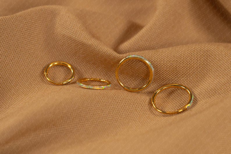 May include: Four gold rings with opal accents are displayed on a tan fabric. The rings vary in size, with one featuring a hinged closure. The opal stones have a subtle, iridescent quality, adding a touch of color.