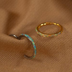 May include: Two rings with opal accents rest on a brown fabric. One ring is gold-toned with a band of opal stones. The other ring is silver-toned with a curved design and opal accents. The rings are likely for the finger.