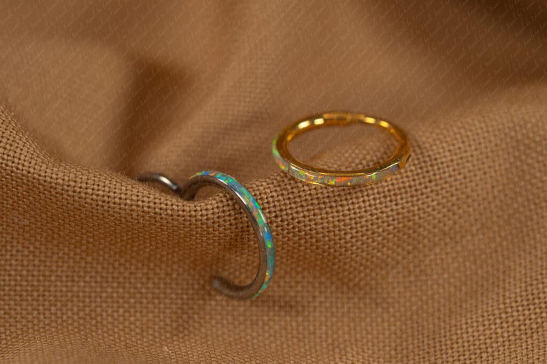 May include: Two rings with opal accents rest on a brown fabric. One ring is gold-toned with a band of opal stones. The other ring is silver-toned with a curved design and opal accents. The rings are likely for the finger.