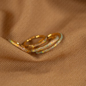 May include: Three gold rings with opal inlays rest on a tan fabric surface. The rings have a delicate, thin band design. The opal inlays display a range of colors, including white, blue, and yellow.