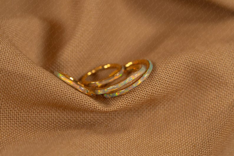 May include: Three gold rings with opal inlays rest on a tan fabric surface. The rings have a delicate, thin band design. The opal inlays display a range of colors, including white, blue, and yellow.