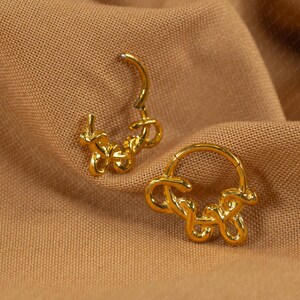 May include: A pair of gold-colored hoop earrings with a unique, abstract design. The earrings are shaped with a looping, flowing style, and are set against a textured, tan fabric background. The earrings are designed to be worn on the ear.