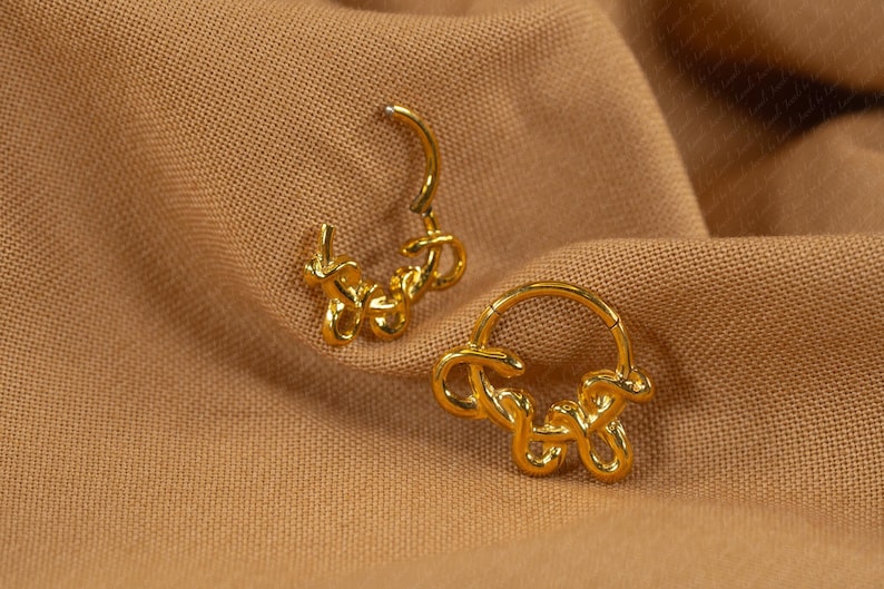 May include: A pair of gold-colored hoop earrings with a unique, abstract design. The earrings are shaped with a looping, flowing style, and are set against a textured, tan fabric background. The earrings are designed to be worn on the ear.