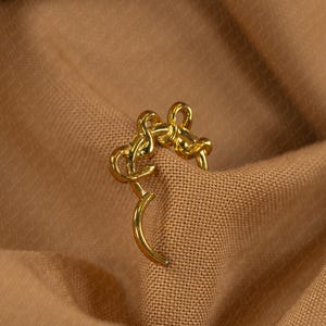 May include: A gold-colored earring with a unique design, featuring a curved post and an intricate, looped upper section. The earring is set against a backdrop of a textured, tan fabric, creating a contrast that highlights its shape and color.