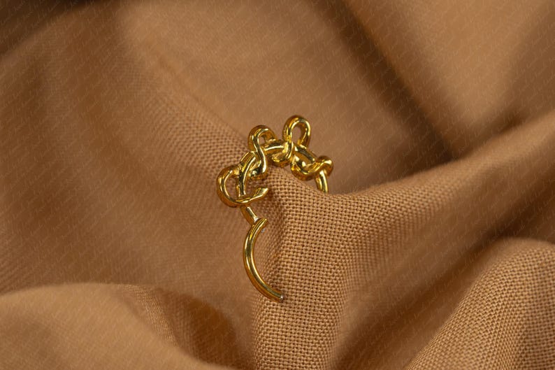 May include: A gold-colored earring with a unique design, featuring a curved post and an intricate, looped upper section. The earring is set against a backdrop of a textured, tan fabric, creating a contrast that highlights its shape and color.