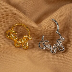May include: Two earrings, one gold and one silver, rest on a tan fabric. The earrings have a looped design, with the gold earring closed and the silver earring open. The earrings are likely for pierced ears.
