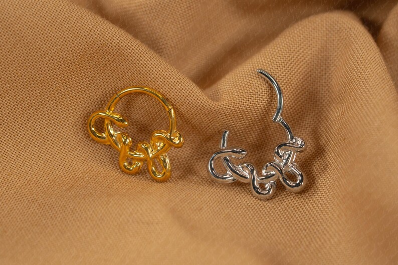 May include: Two earrings, one gold and one silver, rest on a tan fabric. The earrings have a looped design, with the gold earring closed and the silver earring open. The earrings are likely for pierced ears.