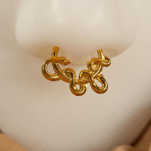 May include: A gold-colored nose ring with a looped design. The jewelry is displayed against a light background, with a hint of a tan fabric in the lower right corner. The nose ring has a unique, intertwined pattern.