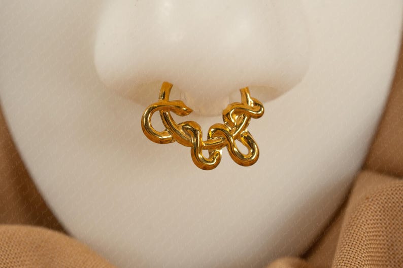May include: A gold-colored nose ring with a looped design. The jewelry is displayed against a light background, with a hint of a tan fabric in the lower right corner. The nose ring has a unique, intertwined pattern.