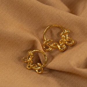 May include: A pair of gold-colored hoop earrings. Each earring features a circular design with an intricate, knotted embellishment. The earrings are displayed on a tan fabric background.
