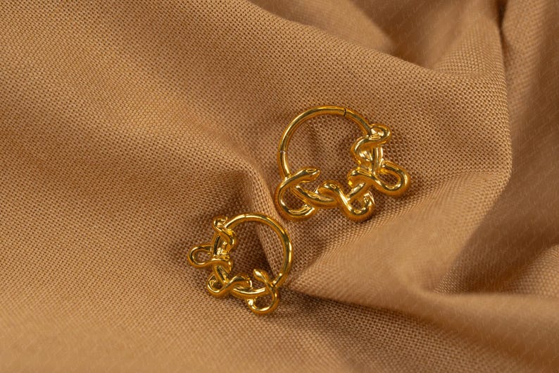May include: A pair of gold-colored hoop earrings. Each earring features a circular design with an intricate, knotted embellishment. The earrings are displayed on a tan fabric background.