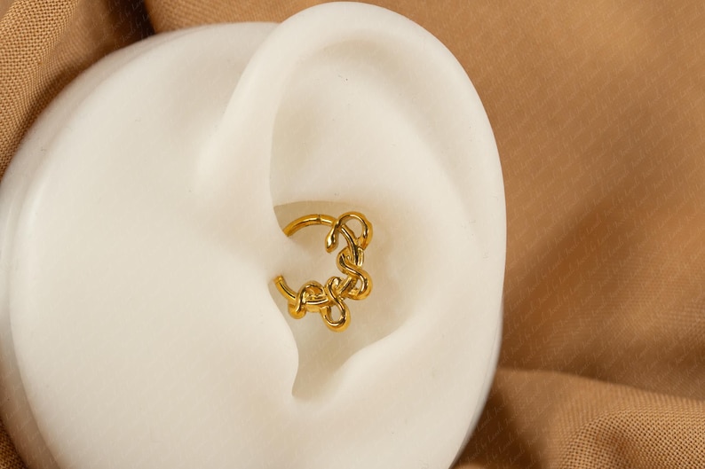 May include: A gold-colored helix earring with a unique, swirling design is shown in the ear. The earring is a hoop style with an intricate, abstract pattern. The ear is set against a neutral background.