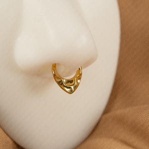 Puede incluir: Un aro de nariz dorado con una superficie texturizada. La joya tiene forma de corazón y se exhibe en una cabeza de maniquí blanca. El fondo es de tela color beige.