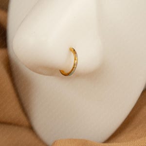 May include: A gold-colored nose ring with a small, curved design. The ring is set against a white background, with a tan fabric in the background. The nose ring is a simple, elegant piece of jewelry.